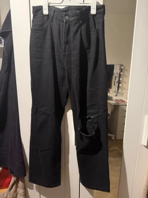 Svarta raka jeans med hål på knät - Säljer ett par svarta jeans med rak passform och stort hål på ena knät. Jeansen har klassisk femficksdesign och hög midja. Perfekta för en avslappnad och trendig look. Materialet är denim i bomull.