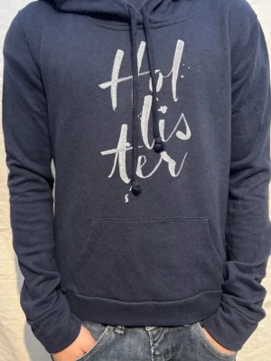 Mörkblå hoodie från Hollister - Mörkblå hoodie från Hollister med stor vit logga framtill. Klassisk känguruficka, justerbar dragsko i huvan och mjukt material som känns skönt mot huden. Perfekt för en avslappnad och trendig stil.