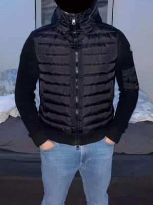 Svart Moncler dunväst med huva - Snygg svart dunväst från Moncler med stickade ärmar och huva. Västen har dragkedja framtill, två sidofickor och en ärmficka med Moncler-logga. Perfekt för lager-på-lager och streetwear-stil.