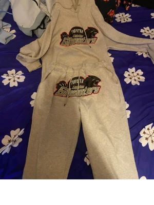 Grå Trapstar Shooters loungewear set - Mysigt grått loungewear set från Trapstar med stor 'Shooters'-logga på både hoodie och byxor. Hoodien har dragsko i huvan och byxorna har resår och dragsko i midjan. Perfekt för chill dagar hemma.