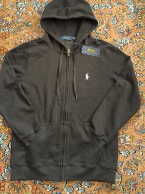 Svart hoodie från Polo Ralph Lauren - Svart hoodie med dragkedja från Polo Ralph Lauren. Klassisk design med huva och dragsko, två fickor framtill och broderad vit logga på bröstet. Mjuk bomullskvalitet som är skön att bära. Perfekt för en avslappnad stil. Sitter bra mellan S-M