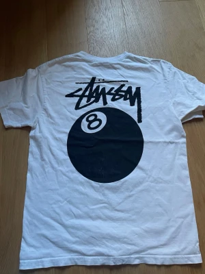 Vit Stüssy t-shirt 8-ball print - Vit t-shirt från Stüssy med stort svart 8-ball tryck och logga på ryggen samt mindre print på bröstet. Klassisk passform och rund hals. Perfekt för dig som gillar streetwear och vill sticka ut med en ikonisk design.