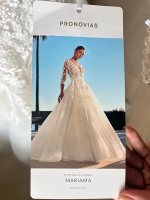Brudklänning från Pronovias - Elegant vit långklänning från Pronovias, modell Mariama. Klänningen har v-ringad front, spetsdetaljer och långärmade ärmar med transparent spets. Kjolen är voluminös och tillverkad i ett lätt, skirt material som ger ett klassiskt och stilrent intryck.