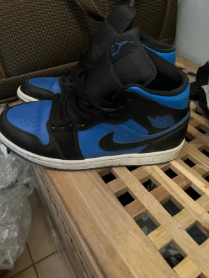 Air Jordan 1 Mid  - Säljer ett par Air Jordan 1 Mid sneakers i svart och blått med klassisk Nike Swoosh på sidan och Air Jordan-logga vid ankeln. Skorna har svart snörning, vit sula och är tillverkade i läder. Perfekta för dig som gillar streetwear och vill sticka ut.