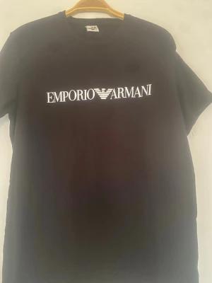 Svart Emporio Armani t-shirt - Svart t-shirt från Emporio Armani med klassisk vit logga tryckt över bröstet. T-shirten har rund halsringning och korta ärmar. Perfekt för en clean och stilren look.