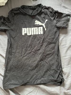 Svart Puma t-shirt med logga - Svart t-shirt från Puma med stor vit logga och text på bröstet. Klassisk rund halsringning och korta ärmar. Tillverkad i mjuk bomull som andas, perfekt för en avslappnad och sportig stil.