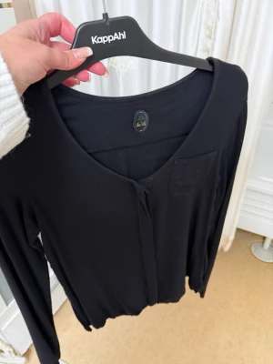 Svart kofta från Nolita med ficka - Svart tunn kofta från Nörda med lång ärm, liten bröstficka och v-ringning. Koftan har en enkel design med diskret logga broderad i guld på ryggen. Perfekt att slänga över en topp för en chill look.