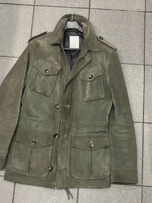 J.lindeberg - Snygg grön mockajacka från .Lindeberg med knappar och dragkedja. Jackan har flera praktiska fickor och axelklaffar för en stilren look. Perfekt för höst och vår. Ny pris 11,499kr