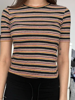 Randig kortärmad topp i bomull - Säljer en kortärmad topp med färgglada horisontella ränder i svart, vit, rosa, gul och orange. Modellen är figurnära och croppad med rund halsringning. Perfekt för dig som gillar en enkel men ändå färgstark stil.