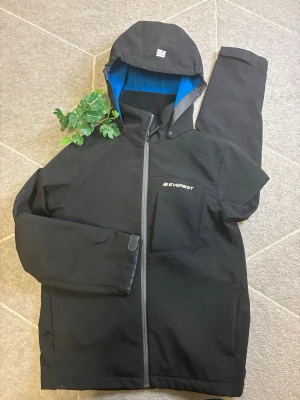 Svart vindjacka från Everest - Snygg svart vindjacka från Everest med blått innerfoder i huvan. Jackan har hel dragkedja framtill, bröstficka med Everest-logga och justerbar huva. Materialet är slitstarkt och vattenavvisande, perfekt för blåsiga dagar. Stilren design med diskreta detaljer.