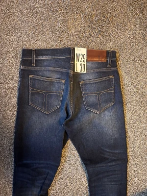 Mörkblå tiger of sweden jeans - Snygga mörkblå jeans med raka ben och klassiska bakfickor. Jeansen har kontrastsömmar och en läderpatch i midjan. Perfekta för dig som gillar en tidlös och clean look. Materialet är troligtvis bomull med stretch för extra komfort.