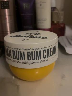 Sol de Janeiro Brazilian Bum Bum Cream - Ikonisk Brazilian Bum Bum Cream från Sol de Janeiro i en rund, gul burk med vitt lock. Innehåller guaranáextrakt och har en fräsch, tropisk doft. Perfekt för dig som vill ha en återfuktande och lyxig kroppskräm.