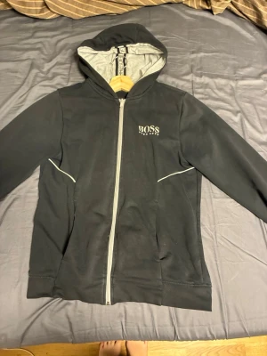 Svart hoodie från Hugo Boss - Snygg svart hoodie från Hugo Boss med dragkedja framtill och vit logga på bröstet. Hoodien har grått innerfoder i huvan och vita detaljer vid fickorna. Perfekt för en chill och stilren look.