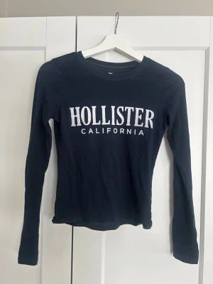 Mörkblå långärmad Hollister topp - Mörkblå långärmad topp från Hollister med vit logga och texten i ”sammet”. Slim fit modell med rund halsringning, tillverkad i mjuk bomull som känns skön mot huden. 