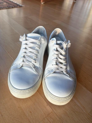 Vita sneakers från Sneaky Steve - Stilrena vita sneakers från Sneaky Steve i läder med klassisk låg profil och vita snören. Skorna har diskret branding på plösen och en chunky vit sula. Perfekta för en clean och fräsch look.