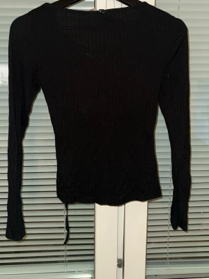 Svart ribbad topp med snörning - Svart långärmad topp i ribbat material med snygg snörning framtill och silverfärgade öljetter. Figurnära passform och stretchigt tyg som ger en cool och trendig look.