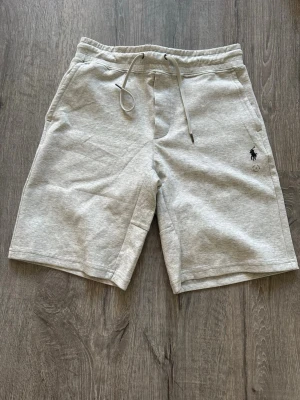 Grå shorts från Polo Ralph Lauren - Snygga ljusgrå shorts från Polo Ralph Lauren med klassisk logga broderad på vänster ben. Mjukt bomullsmaterial, elastisk midja med snörning och två sidofickor. Perfekta för en avslappnad och stilren look.