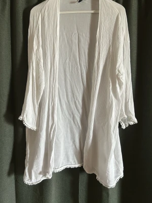 Vit öppen cardigan från Divided - Luftig vit cardigan från Divided med öppen front och trekvartsärmar. Materialet är tunt och lätt strukturerat, och kanterna är dekorerade med små bollar för en söt touch. Perfekt att slänga över en topp under varmare dagar.