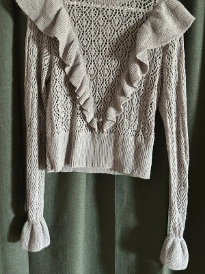 Beige stickad tröja med volang - Supersöt beige stickad tröja med djup V-ringning och volangdetaljer längs halsen. Tröjan har ett genombrutet spetsmönster och långa ärmar med breda muddar. Perfekt för dig som gillar romantisk och trendig stil.