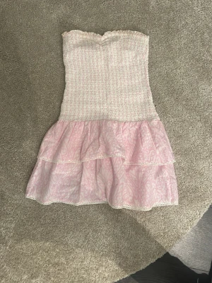 Rosa axelbandslös klänning Gina Tricot - Söt axelbandslös kortklänning från Gina Tricot i ljusrosa och vitt. Klänningen har smock upptill, volangkjol i två lager och diskret mönster. Perfekt för sommarens alla tillfällen!