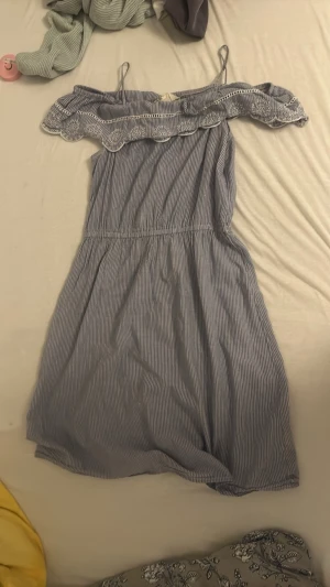 Blåvit offshoulder sommarklänning - Söt blåvit randig offshoulder klänning med smala axelband och volang upptill. Klänningen har resår i midjan och är tillverkad i ett lätt bomullstyg, perfekt för varma dagar. Volangdetaljen har spetsmönster för extra somrig känsla.