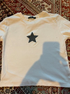 Vit t-shirt med svart stjärna - Vit t-shirt/magtröja från Ambar med en svart stjärna tryckt mitt på bröstet. Klassisk rund halsringning och korta ärmar. Perfekt för en enkel och stilren look.