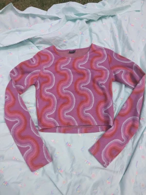 Lila och rosa mönstrad croppad topp - Säljer en croppad långärmad topp från Gina Tricot i lila och rosa med vågigt mönster i vitt och rött. Toppen har rund halsringning! Perfekt för dig som gillar färg och retrovibbar!