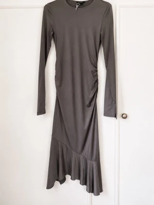 Grå långärmad långklänning - Snygg grå långklänning med lång ärm och rund halsringning. Klänningen har en asymmetrisk nederkant och lätt rynkning i sidan som ger en cool detalj. Tillverkad i ett mjukt, stretchigt material som sitter bekvämt på kroppen.