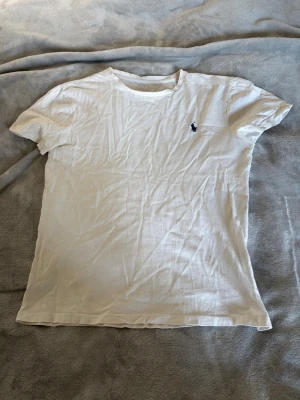 Vit t-shirt från Polo Ralph Lauren - Klassisk vit t-shirt från Polo Ralph Lauren med den ikoniska blå loggan broderad på bröstet. T-shirten har rund halsringning och korta ärmar. Tillverkad i mjuk bomull för en skön och avslappnad stil.