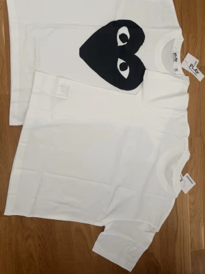 Vita Comme des Garçons PLAY t-shirts - Två vita t-shirts från Comme des Garçons PLAY. Ena t-shirten har det ikoniska svarta hjärta-trycket med ögon på bröstet, medan den andra är helt vit. Båda har rund hals och korta ärmar. Perfekta för en clean och stilren look.