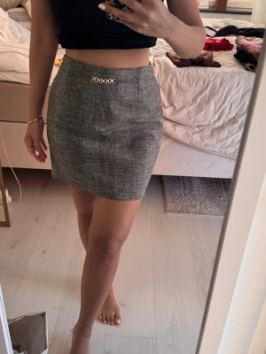 Grå rutig kortkjol med kedjedetalj - Snygg grå rutig kortkjol med en liten guldfärgad kedjedetalj framtill i midjan. Kjolen har en klassisk rak passform och stängs med dragkedja bak. Perfekt att styla med en crop top eller stickad tröja.