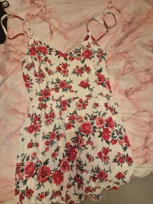 Vit blommig sommarklänning med rosor - Söt vit klänning med röda rosor och gröna blad, smala axelband och v-ringad hals. Klänningen har en kort längd och är perfekt för varma dagar. Tillverkad i lätt bomullsmaterial som känns skönt mot huden. Passformen är normal och modellen är luftig och bekväm.