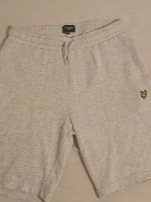 Grå sweatshorts från Lyle & Scott - Mysiga ljusgrå sweatshorts från Lyle & Scott med elastisk midja och snörning. Klassisk gul logga på vänster ben och en bakficka. Perfekta för chill dagar eller träning.