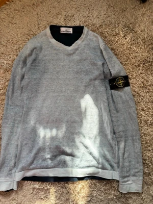 Ljusgrå stickad tröja Stone Island - Snygg ljusgrå stickad tröja från Stone Island med klassisk svart och gul logotyp-patch på vänster ärm. Tröjan har rund halsringning och långa ärmar. Perfekt för dig som gillar stilrena plagg med streetwear-vibe. 2 i 1 tröja, vänd den ut och in för att få en mörkblå tröja. I fint skick. 