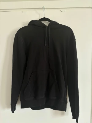 Svart hoodie med magficka - En klassisk svart hoodie med huva och dragsnören. Hoodien har en stor magficka framtill och ribbade muddar vid ärmslut och nederkant. Perfekt för en avslappnad och stilren look. 
