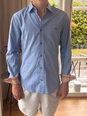 Burberry skjorta  - Burberry skjorta – Strl S  Säljer en Burberry-skjorta i storlek S. Ljusblå modell med broderad Burberry-logga på bröstet och klassiskt Burberry-rutmönster på insidan av ärmsluten. Superbra skick, sparsamt använd och utan fläckar eller hål.  ✅ Storlek: S ✅ Superbra skick ✅ Snabb frakt 📦  Hör gärna av dig vid frågor eller intresse!