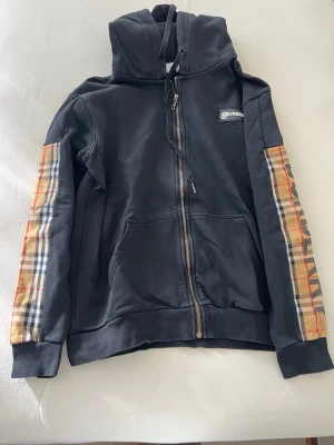 Svart Burberry hoodie med rutiga ärmar - Snygg svart hoodie från Burberry med dragkedja och huva. Ärmarna har klassiskt Burberry-rutigt tyg i beige, svart, vitt och rött samt stor logga längs ena ärmen. Mjuk bomullsmix och ribbade muddar. Perfekt för en trendig streetwear-look.