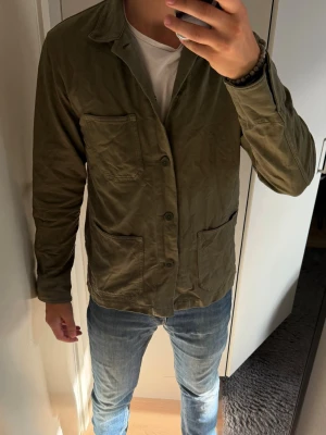 Olivgrön overshirt med fickor - Snygg olivgrön overshirt med tre stora utanpåliggande fickor och knäppning framtill. Strl S men sitter som M. Overshirten har en avslappnad passform, klassisk krage och är tillverkad i ett slitstarkt bomullstyg. Perfekt att bära över en t-shirt för en casual look.