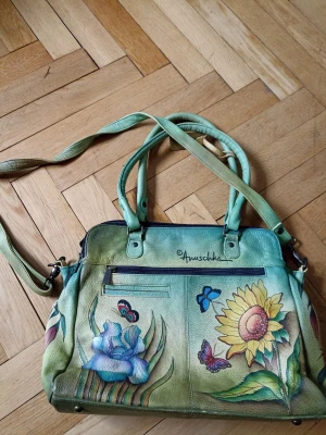 Grön handväska med blommor från Anuschka - Unik handväska i grönt läder från Anuschka med handmålade blommor, fjärilar och bin i färgglada nyanser. Väskan har både handtag och axelrem samt flera fack med dragkedja. Perfekt för dig som gillar konstnärliga detaljer och naturmotiv.