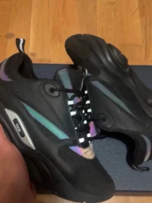 Svarta sneakers med färgdetaljer - Säljer ett par svarta Puma sneakers med chunky sula och coola iriserande detaljer i lila, blått och grönt. Skorna har svart mesh och syntetmaterial, samt svartvita snören för extra stilpoäng. Perfekta för dig som vill sticka ut med dina skor.