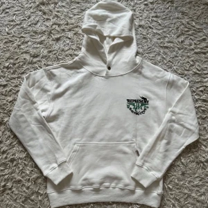 Represent- hoodie - Snygg vit hoodie från Represent med tryck i grönt och svart på bröstet och ryggen där det står 'Represent Racing Club'. Hoodien har huva, magficka och ribbade muddar. Perfekt för dig som gillar streetwear och vill sticka ut med en unik design.