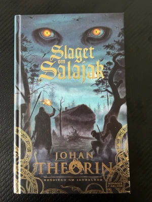 Slaget om Salajak av Johan Theorin - Fantasybok med hård pärm, mörkblå och svart omslag med gyllene detaljer och mystiska illustrationer. Första delen i Krönikan om Jarmaland, skriven av Johan Theorin. Perfekt för dig som gillar äventyr, magi och nordisk fantasy.