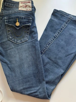 Blå True Religion jeans med fickdetaljer - Säljer ett par klassiska blå jeans från True Religion med ikoniska kontrastsömmar och stora fickor bak med lock och guldfärgade knappar. Jeansen är bootcut. De är i storlek 24 men uppsydda en liten bit. Jag är 165. Perfekt för dig som gillar low Rise bootcut jeans. De är nya utan prislapp.