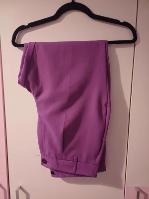 Byxor - Snygga lila kostymbyxor med hög midja och raka ben. Byxorna har fickor i sidorna och stängs med knapp och dragkedja. Perfekta för dig som vill sticka ut med färg och ha en trendig look. Materialet känns mjukt och följsamt.