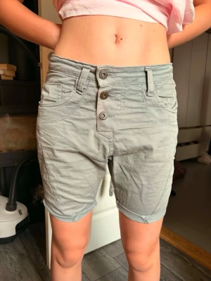 Gråa high waist shorts från Please - Säljer ett par gråa högmidjade shorts från Please med tre knappar framtill och klassiska fickor både fram och bak. Shortsen har en avslappnad passform och är tillverkade i bomull, perfekta för varma dagar.