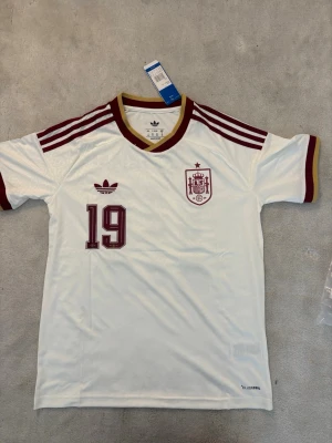 Vit Spanien fotbollströja Adidas storlek S och M - Säljer en vit fotbollströja från Adidas med Spaniens landslagsemblem och nummer 19 i vinrött. Tröjan har vinröda detaljer på axlar och ärmslut samt guldfärgad kant vid halsen. Tillverkad i lätt och ventilerande material, perfekt för träning eller match.