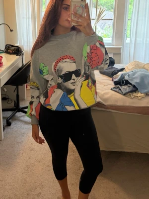 Grå hoodie med färgglad pop art-print - Säljer en grå hoodie med oversized passform och färgglad pop art-inspirerad print på både fram- och baksida. Motivet visar ansikten med solglasögon och starka färger som rött, blått, gult och grönt. Hoodien har ribbade muddar och är perfekt för dig som vill sticka ut.