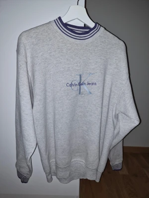Grå sweatshirt från Calvin Klein Jeans - Ljusgrå sweatshirt med broderad CK-logga och Calvin Klein Jeans-text på bröstet. Tröjan har ribbade muddar och krage med lila och vita ränder. Tillverkad i mjukt material och har en relaxed fit som ger en clean och stilren vibe.