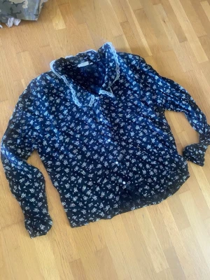 Vintage blus med spetskrage - Söt mörkblå blus med vitt blommönster och dekorativ spetskrage. Blusen har axelvaddar långa ärmar och knäppning framtill. Perfekt för dig som gillar romantisk och vintageinspirerad stil.