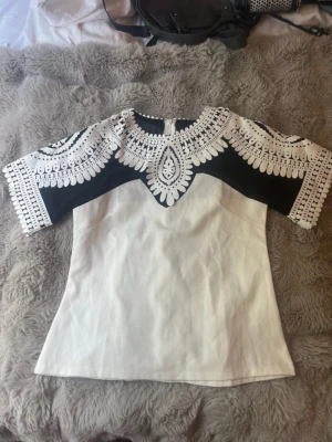 Vit och svart blus med mönster - Säljer en unik blus i vitt och svart med korta ärmar och ett detaljerat grafiskt mönster upptill. Blusen har rund halsringning och är tillverkad i ett fast tyg som ger en snygg passform.